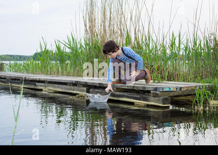 Un ragazzo su una banchina in legno osserva barche di carta. Foto Stock