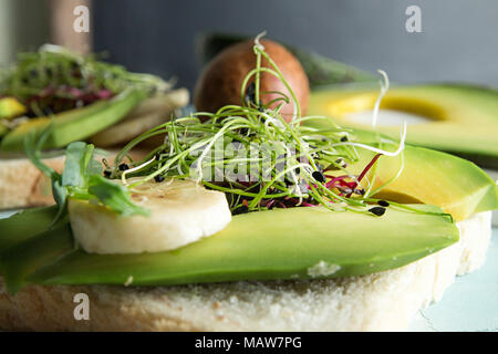 Sana colazione toast con crema di formaggio, fette di avocado, banana e microgreen su sfondo di calcestruzzo. Pulire mangiando la dieta vegana concetto. Chiudere Foto Stock