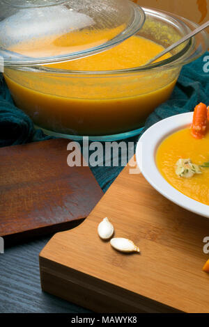 Calda fresca zuppa di zucca nel piatto di vetro e la piastra bianca, decorata con mini carota, semi di zucca e a forma di fungo presente sul pannello di legno Foto Stock