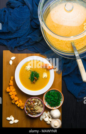 Calda zuppa di zucca nel piatto di vetro e la piastra bianca, decorata con mini carota, semi di zucca, funghi, miele e agaric kelp verde sul pannello di legno. Ve Foto Stock