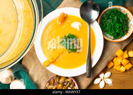 Calda zuppa di zucca nel piatto di vetro e la piastra bianca, decorata con mini carota, semi di zucca, funghi, miele e agaric kelp verde sul pannello di legno. Ve Foto Stock