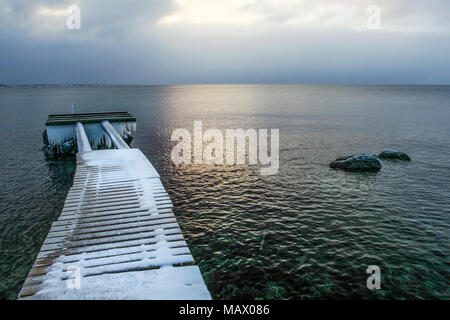 La barca di legno pier coperto di neve e ghiaccio con ghiaccioli alla fine, con il sole che splende sul mare in background. Foto Stock