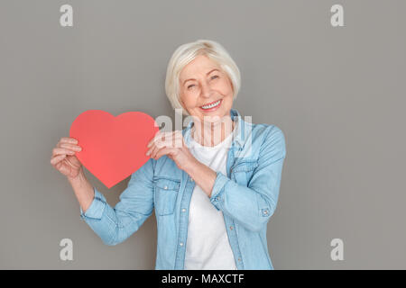 Le donne più anziane indossano jeans camicia studio isolato su sfondo grigio per celebrare il giorno di san valentino tenendo la carta rossa cuore predisposto alla ricerca della fotocamera Foto Stock