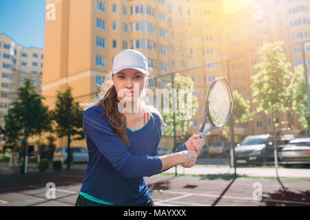 La donna , guardando la fotocamera e azienda racchetta da tennis. Attraente giocatore di tennis giocando a tennis all'aperto,praticando sulla corte alla giornata di sole su una città b Foto Stock