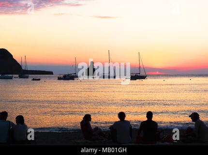 Tramonto di scena a Spiaggia Benirras, Isola di Ibiza. Formazione di roccia e sunbeam, vacanze estate scena. La gente a guardare il tramonto sulla spiaggia. Foto Stock