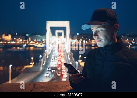 Giovane uomo nella città di controllo telefono mobile lungo la strada di notte. Budapest, Ungheria. Foto Stock