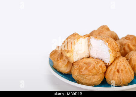 Close up Profiteroles freschi bignè torte ripiene con crema pasticcera e con uno tagliate a metà in un turchese piastra blu su sfondo bianco. E Foto Stock