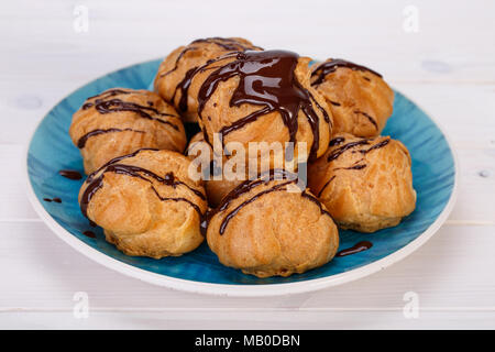 Profiteroles freschi bignè torte ripiene con crema pasticcera e guarnita con salsa al cioccolato in un turchese piastra blu su sfondo bianco.popolari P Foto Stock