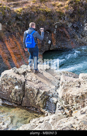 Uomo in piedi sulle rocce azienda panasonic videocamera per catturare il paesaggio alla Fairy Piscine, fiume fragile, Isola di Skye in Scozia, nel Regno Unito nel mese di marzo Foto Stock