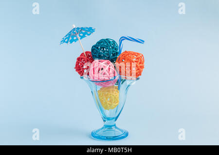 Gelato il gelato con colorati gelato palline fatte di sfere di rattan, ombrello di carta e cannuccia in coppa contro pastello blu sullo sfondo c minimo Foto Stock