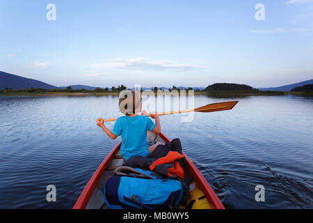 Ragazzo giovane paddling in una canoa su un lago al crepuscolo Foto Stock