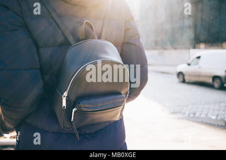 Vista posteriore della donna con zaino in pelle indossando down jacket va sulla strada della citta' al mattino o alla sera. Raggi di sole risplendere. Dettagli del casual abito invernale. Foto Stock