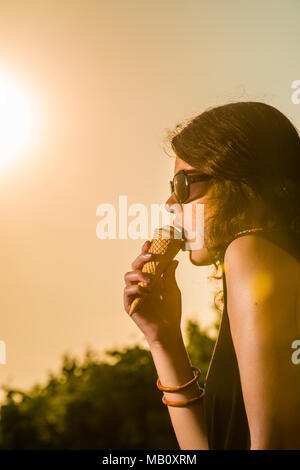 Giovane e attraente donna mangiare gelato nel verde durante il tramonto con occhiali da sole Foto Stock