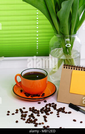 Tulipani gialli in un vaso, un arancione con la tazza di caffè nero, i chicchi di caffè, una penna e un blocco note stand sul davanzale. La molla ancora vita Foto Stock