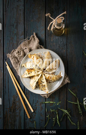 Asian Gyozas gnocco fritto potstickers su piastra in ceramica, servita con bacchette e bottiglia di salsa sul legno scuro plank sfondo. Vista superiore, spa Foto Stock