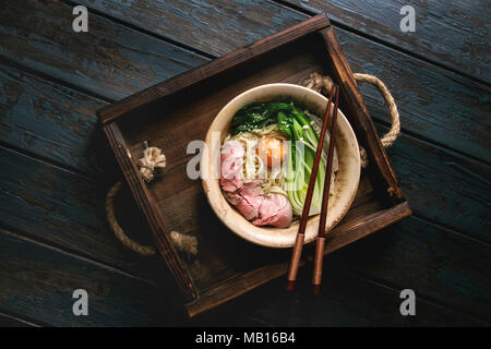 Piatto asiatico udon tagliatelle con tuorlo d'uovo, sesamo, funghi, boc choy, affettato sous vide carne cotta servito in tazza ceramica con cucchiaio e bastoncini su w Foto Stock