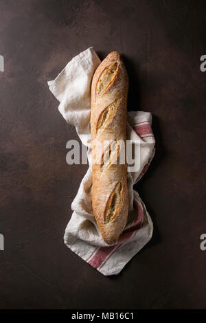 Filone di freschi di forno artigiano baguette di pane sulla tela di lino su marrone scuro dello sfondo texture. Vista superiore, copia dello spazio. Foto Stock