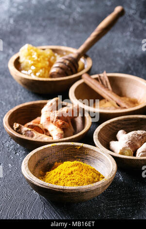 Ingredienti per la curcuma latte. Curcuma in polvere, curcuma radice, la cannella, lo zenzero, i favi in ciotole di legno nero su sfondo di texture. Close up Foto Stock