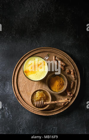 Tazza di bevanda ayurvedica golden latte curcuma latte con curcuma in polvere il round vassoio in legno e gli ingredienti di cui sopra nero su sfondo di texture. Top vi Foto Stock