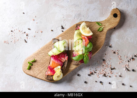 La barbabietola rossa a fette di salmone marinato panini con pane di segale, cetriolo, basilico e limone servita sul tagliere di legno su grigio Sfondo texture. Parte superiore Foto Stock