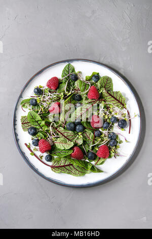 Vegano verde insalata con frutti di bosco fragola, mirtillo, germogli giovani foglie di bietole su piastra in ceramica su cemento grigio Sfondo. Vista superiore, spazio. Foto Stock