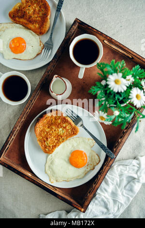 Facile toast alla francese servita con un uovo fritto sul lato su una piastra bianca, una tazza di caffè e latte e fiori di primavera in un vassoio di legno fotografato da a Foto Stock