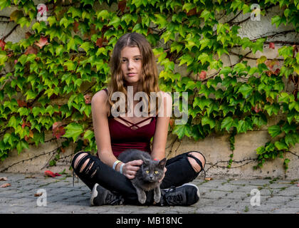 Caucasico bella ragazza adolescente siede in giardino con il suo gatto di casa. Pretty girl teen, estate, top, siede zampe trasversale di coccole carino gattino grigio, all'esterno. Foto Stock