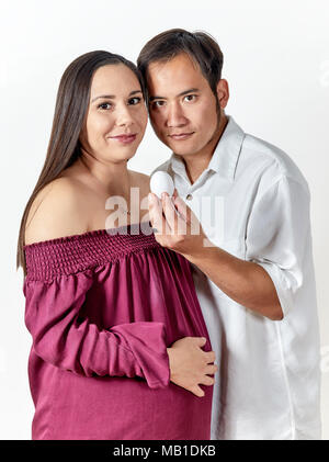 Attraente giovane donna incinta con il suo partner di sorridere mentre tiene un uovo e guardando la telecamera isolato su bianco Foto Stock