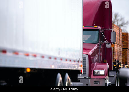 Big Rig potente classico rosso scuro semi carrello trattore il trasporto di pacchi di pannelli di legno legname sul letto piano semi rimorchio la guida in convoglio dietro anoth Foto Stock