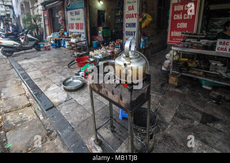 Hanoi, Vietnam - 24 Gennaio 2016: un proprietario di un negozio di mettere un bollitore grande sul braciere di un contenitore per il carbone caldo per riscaldare per fare il tè in un affollato centro Quart Foto Stock