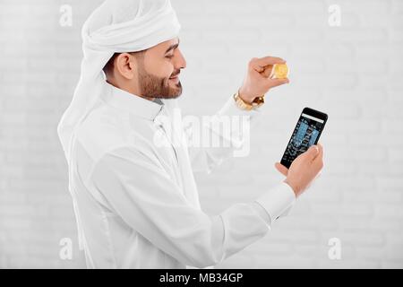 Giovani Musulmani trader continua medaglia d'oro e telefono cellulare in un'altra mano. Egli si erge sul bianco di sfondo per studio. Un uomo guarda felice e impegnata. Foto Stock