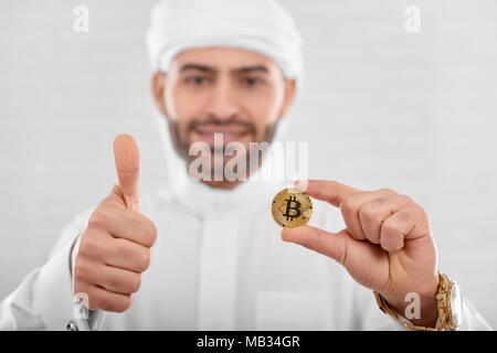 Giovani Musulmani trader continua golden coin sulla bianca di sfondo per studio. Un uomo guarda felice e impegnata. Egli indossa tradizionale costume arabo e sembra soddisfatta. Foto Stock