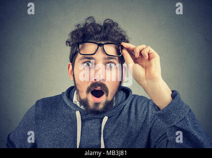 Giovani barba casual uomo prendendo il largo occhiali e guardando la fotocamera con shock. Foto Stock