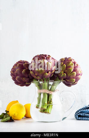 Mazzetto fresco di viola i carciofi in un decantatore con acqua su sfondo bianco con limone, olio di oliva , aglio . molla cibo sano Foto Stock