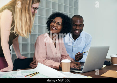 Gruppo di colleghi diverso sorridente e avente una discussione su un laptop mentre si lavora insieme a una scrivania in un ufficio moderno