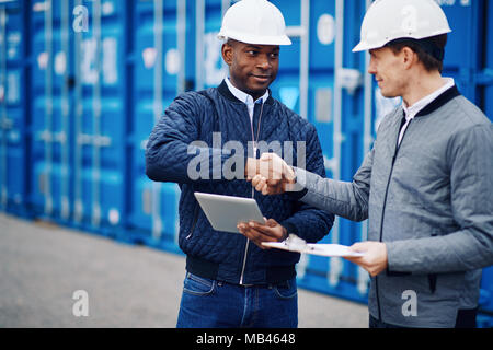 Due ingegneri sorridente indossando hardhats stringono le mani insieme da contenitori di trasporto merci su un dock commerciale Foto Stock