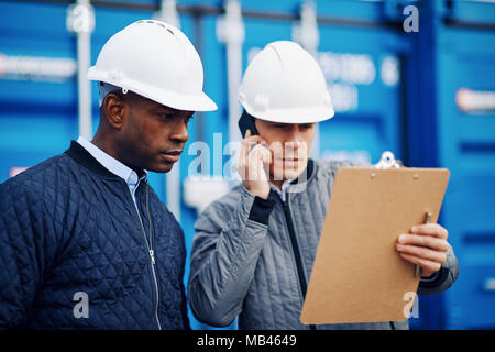 Gestore del porto in piedi con un collega in una spedizione commerciale cantiere parlando al cellulare e la lettura di un elenco inventario sulla clipboard Foto Stock