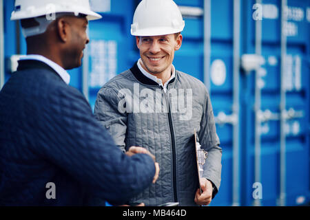 Due ingegneri sorridente indossando hardhats in piedi da contenitori di trasporto merci su un dock commerciale agitando mani insieme Foto Stock