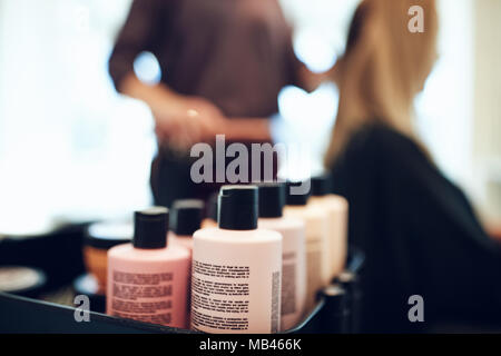 Primo piano di shampoo e balsamo bottiglie su un vassoio in un salone di bellezza con parrucchiere styling i capelli dei clienti in background Foto Stock