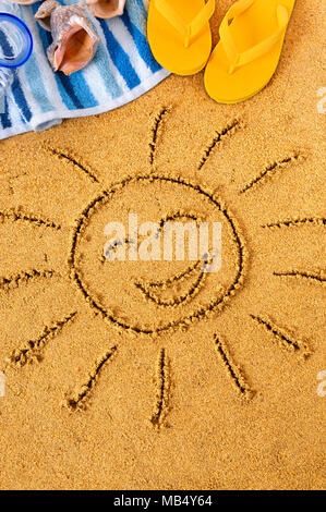 Smiley face sun disegnato in sabbia su una spiaggia con asciugamano, conchiglie e flip flop Foto Stock
