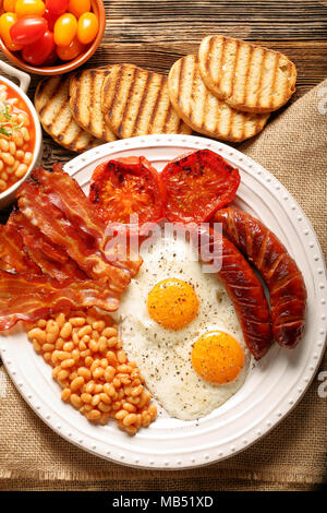 Prima colazione inglese con salsicce, pomodori grigliati, uova, pancetta, fagioli e pane sulla piastra bianca Foto Stock