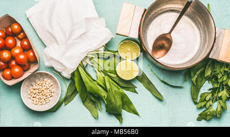 Aglio selvatico la cottura di preparazione: pot con cucchiaio, aglio selvatico, pinoli, pomodori e condire su sfondo blu, vista dall'alto. Una sana cucina di stagione un Foto Stock