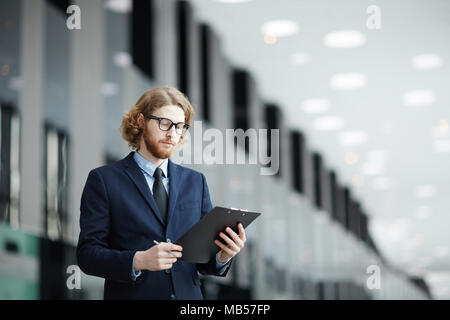 Giovane imprenditore della tuta e occhiali lettura documento finanziario nella lounge del moderno aeroporto Foto Stock