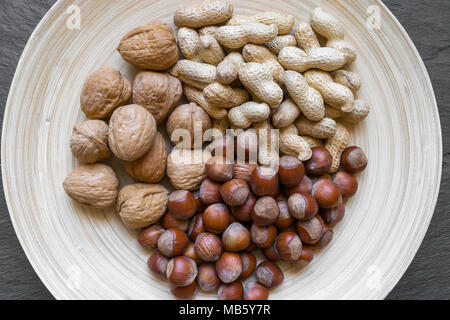 Arachidi, nocciole e noci con nutshells mix in bamboo naturale della piastra sulla pietra nera superficie di sfondo Foto Stock