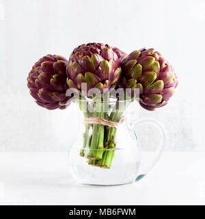 Mazzetto fresco di viola i carciofi in un decantatore con acqua su sfondo bianco . molla cibo sano con spazio di copia Foto Stock