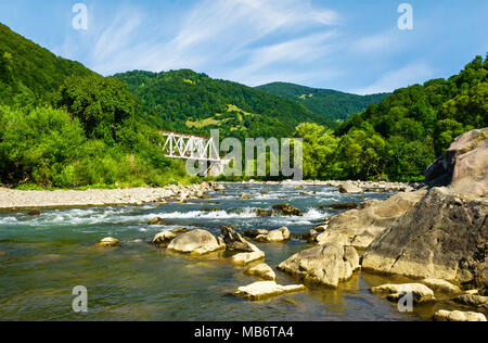 Fiume nelle montagne boscose. bellissimo paesaggio estivo con enormi rocce sulla riva e un metallo bianco ponte di treno di distanza Foto Stock