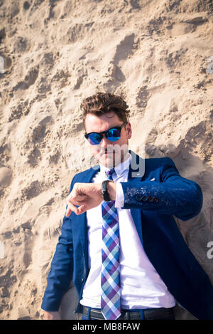 Funny manager perse nel tempo ma rimane calmo. Giovane serio in elegante abito è in ritardo sul lavoro. Elegante ragazzo giace sulla spiaggia e si affaccia sull'orologio Foto Stock