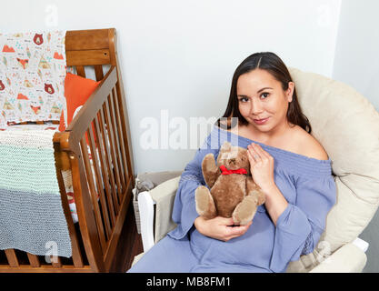 Attraente donna incinta seduto in una sedia a dondolo accanto a una culla per neonati tenendo un orsacchiotto di peluche e sorridente Foto Stock