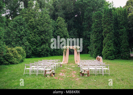 Bellissimo luogo reso con piazza di legno e rose floreali decorazioni per esterno cerimonia di matrimonio nel parco verde. Righe di tante vuote con sedie in legno leggere Foto Stock