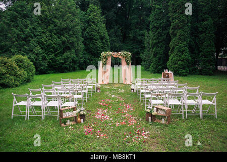 Bellissimo luogo reso con piazza di legno e rose floreali decorazioni per esterno cerimonia di matrimonio nel parco verde. Righe di tante vuote con sedie in legno leggere Foto Stock